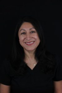Professional headshot of a smiling woman with dark, shoulder-length hair wearing a black top and a delicate pendant necklace against a plain black background