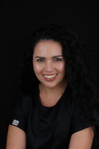 Professional headshot of a smiling woman with long curly black hair, wearing a black top, posing against a solid black background.